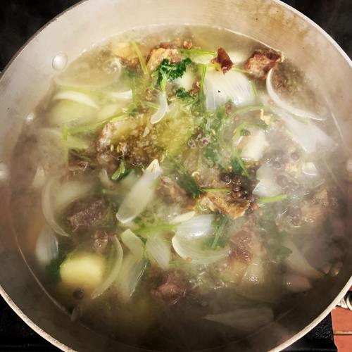 A pot of simmering soup with meat, onions, potatoes, and herbs, creating steam on a stovetop.
