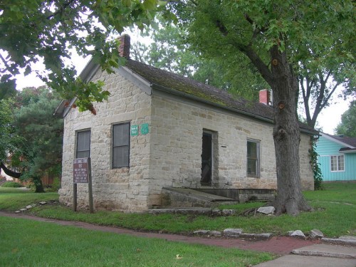 Historic stone house with a wooden porch, surrounded by trees and a grassy area, featuring a nearby informational sign.