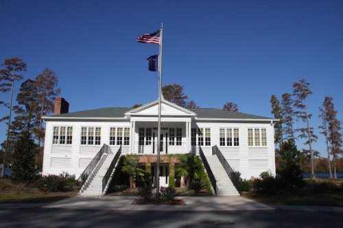 A white building with a flagpole in front, surrounded by trees and a clear blue sky. Steps lead up to the entrance.