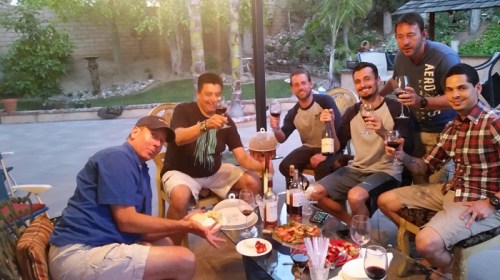 A group of six men enjoying a gathering outdoors with food, drinks, and laughter in a backyard setting.