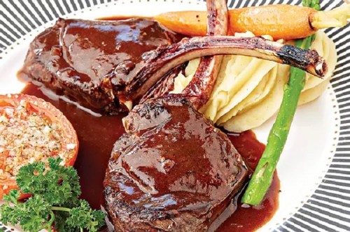 A plate featuring two lamb chops with rich sauce, mashed potatoes, carrots, asparagus, and a stuffed tomato.