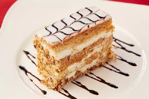 A slice of layered pastry dessert with cream and chocolate drizzle on a white plate.