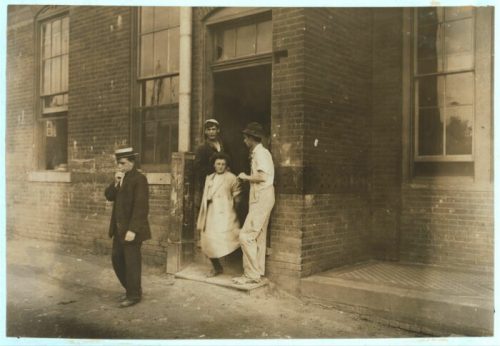 A group of children, including a girl in a dress, exit a brick building while a boy in a hat stands nearby.