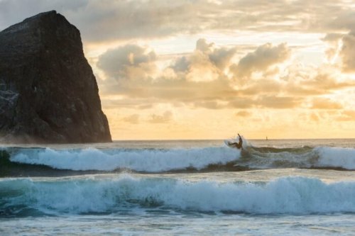 A surfer rides a wave at sunset, with a rocky cliff in the background and clouds illuminated by warm light.