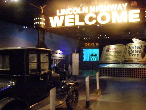A vintage car in front of a "Welcome" sign for the Lincoln Highway, with a large open book display in the background.