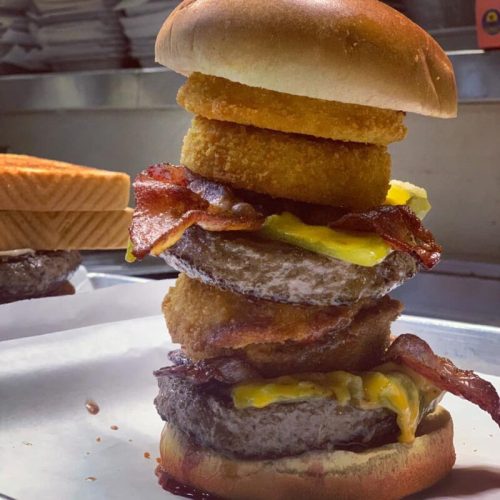 A towering burger with two beef patties, cheese, bacon, onion rings, and a top bun, served on a white paper.