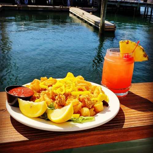 A plate of fried calamari with lemon and dipping sauce, alongside a colorful cocktail, by the water.