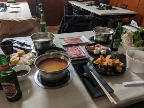 A table set for hot pot with various ingredients, including meats, vegetables, and drinks, in a restaurant setting.