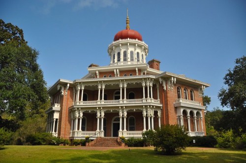 A grand, historic mansion with a red dome, white columns, and lush greenery surrounding it under a clear blue sky.