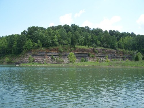A serene lake with a rocky shoreline and lush green trees under a clear blue sky.