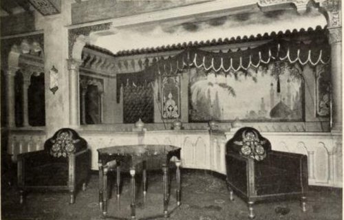 A vintage interior featuring ornate chairs, a decorative table, and a mural backdrop in an elegant setting.
