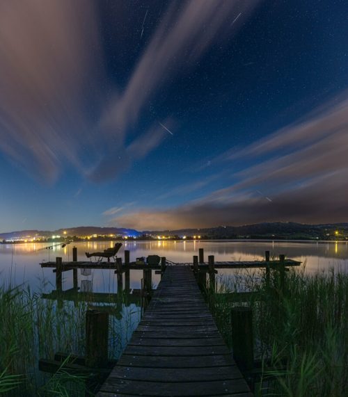 A serene lakeside scene at night, featuring a wooden dock, starry sky, and soft clouds reflecting on the water.