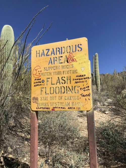 Sign warning of hazardous area with notes about slippery rocks, flash flooding, and safety precautions. Cacti in background.