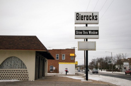 Sign for "Bierocks" with a drive-thru window, snowy street scene, and a person walking in the background.