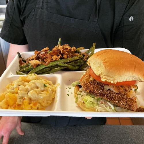 A person holds a food tray with a fried chicken sandwich, macaroni and cheese, green beans, and crispy onions.