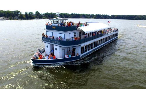 A large boat named "Madam Carroll" with passengers on deck cruising on a lake under a clear sky.