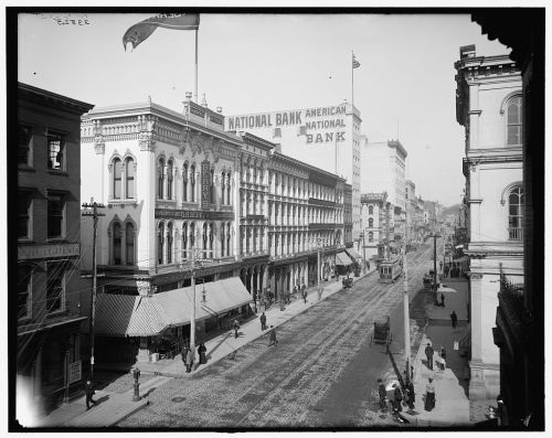 Historic street view featuring the National Bank building, shops, and a streetcar in a bustling urban setting.