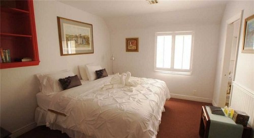 Cozy bedroom with a large bed, white linens, a lamp, and a window, featuring light-colored walls and red accents.
