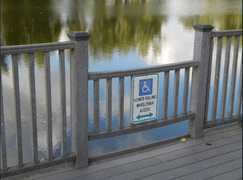 Sign on a wooden railing by a pond indicating wheelchair access with an arrow pointing left.