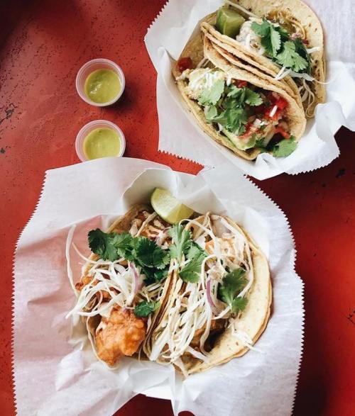 Two tacos filled with fresh ingredients, garnished with cilantro, served with lime and small cups of green sauce.