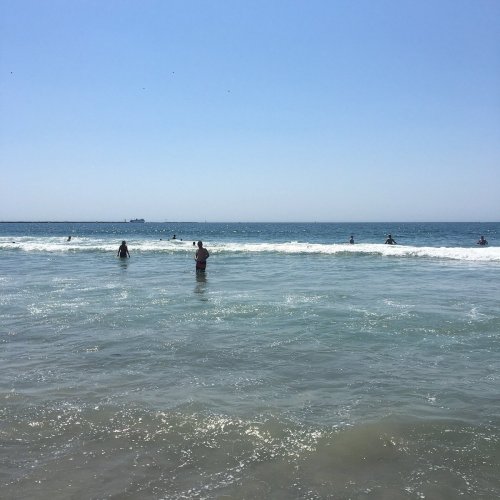 People wade in the ocean waves on a sunny day, with a clear blue sky and distant boats on the horizon.