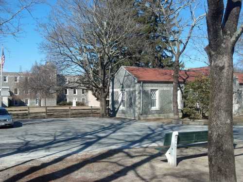 A quiet park scene featuring a gray building, trees, and a bench, with a car parked nearby on a gravel path.