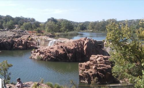 A serene landscape featuring a rocky shoreline, a small waterfall, and a calm lake surrounded by greenery.