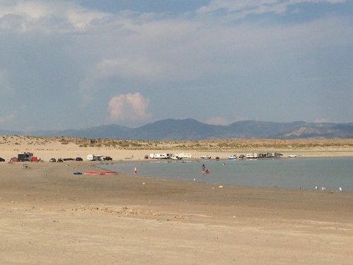 A sandy beach with vehicles and campers near a calm lake, surrounded by mountains under a cloudy sky.