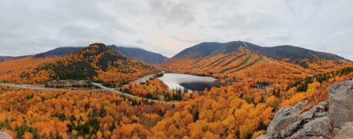 A panoramic view of a vibrant autumn landscape with orange and yellow trees surrounding a calm lake and mountains.