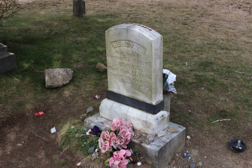 A weathered gravestone with the name "Mercy" and flowers at its base, set in a grassy cemetery.