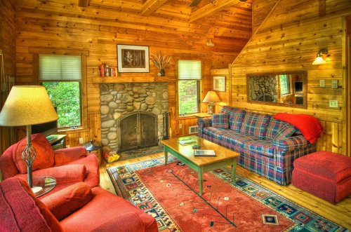 Cozy wooden cabin living room with a stone fireplace, plaid sofas, a coffee table, and a colorful rug.