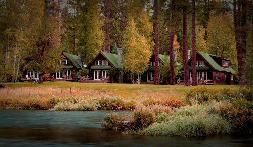 A serene landscape featuring rustic cabins by a river, surrounded by lush trees and autumn foliage.