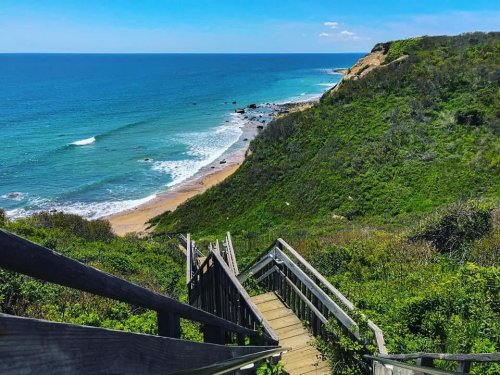 A wooden staircase leads down to a sandy beach surrounded by lush greenery and a calm blue ocean.