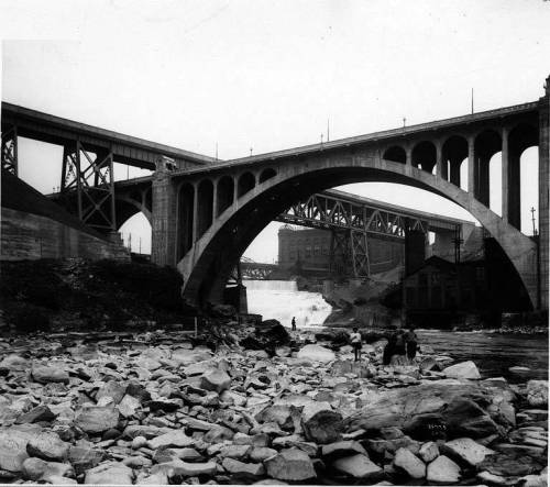 Historic black-and-white photo of bridges over a river, with rocky banks and a waterfall in the background.