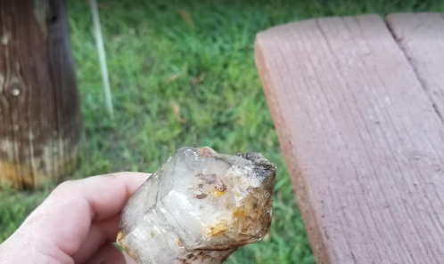 A hand holding a large, translucent crystal with a rough surface, set against a grassy background and wooden surface.