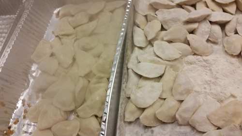 Two trays of uncooked dumplings, one in a container and the other laid out on a floured surface.