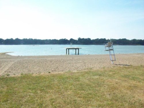 A sandy beach with a wooden dock and a lifeguard chair overlooking a calm lake under a clear sky.