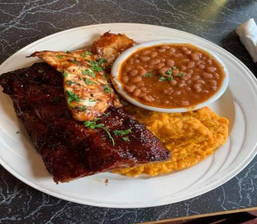 A plate featuring BBQ ribs, grilled chicken, baked beans, and mashed sweet potatoes, garnished with parsley.