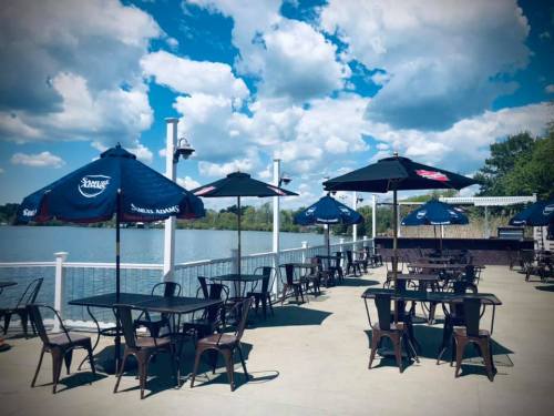Outdoor dining area by a lake, featuring tables and umbrellas under a bright blue sky with fluffy clouds.