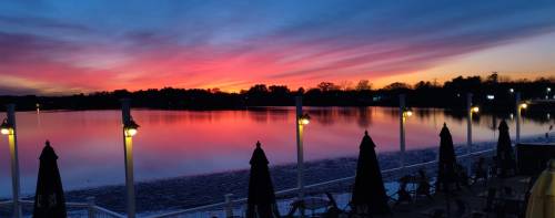 A vibrant sunset over a calm lake, with silhouetted umbrellas and lights reflecting on the water.