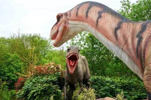 Two large dinosaur sculptures with open mouths, surrounded by greenery and trees in a natural setting.