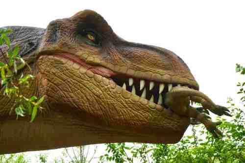 A large dinosaur sculpture, resembling a T-Rex, holds a smaller dinosaur in its mouth amidst green foliage.
