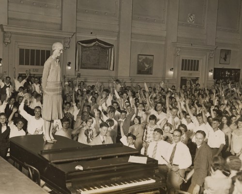 A crowd of people enthusiastically raises their arms as a woman stands on a piano, addressing them in a large hall.