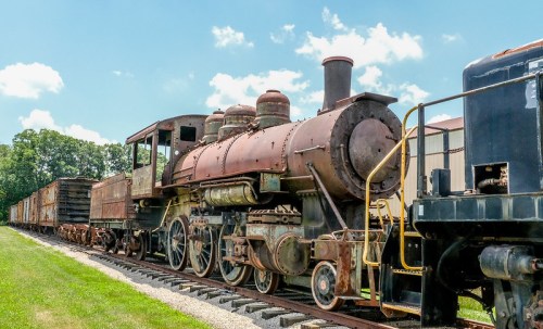 Rusty vintage steam locomotive parked on tracks, surrounded by old freight cars under a blue sky with fluffy clouds.