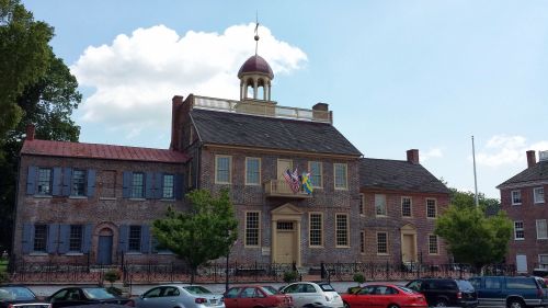 Historic brick building with a dome, flags, and trees, set against a blue sky with clouds. Cars parked in front.
