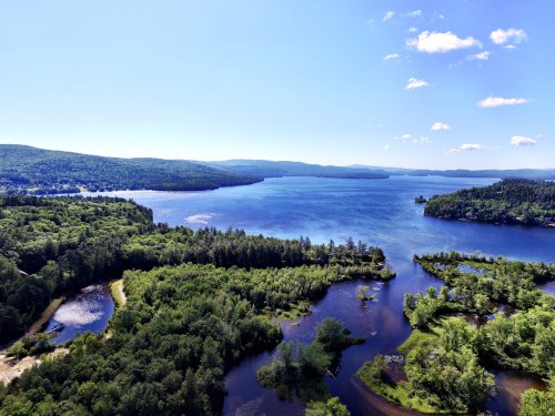 Aerial view of a serene lake surrounded by lush green forests and rolling hills under a clear blue sky.