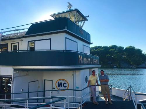 Two men stand beside the "Madam Carol" boat, with a calm water backdrop and clear blue sky.