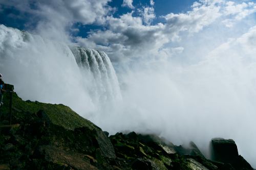 A powerful waterfall cascades over rocky cliffs, surrounded by mist and dramatic clouds in the sky.