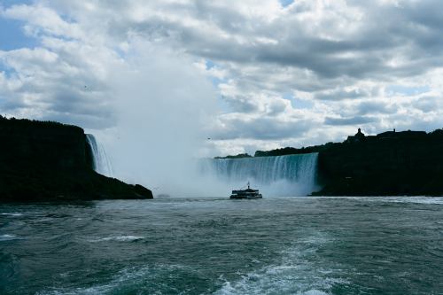 A boat sails near Niagara Falls, with mist rising and clouds overhead, capturing the natural beauty of the scene.