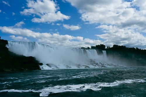 A stunning view of a waterfall cascading over cliffs, surrounded by mist and a blue sky with fluffy clouds.